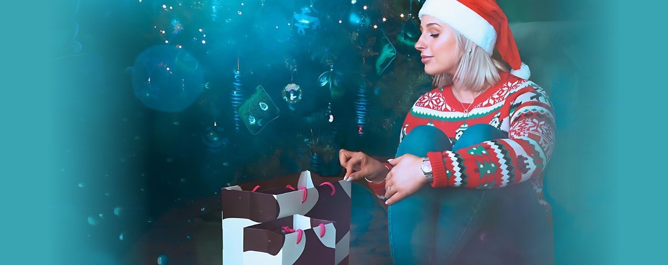 Lady in Christmas jumper sat in front of Christmas tree peeping in gift bag