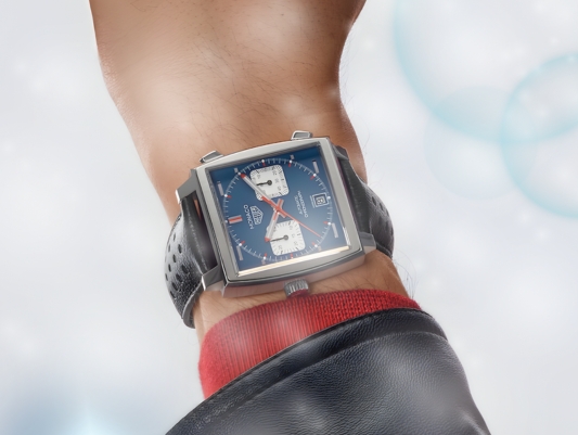 A man wearing a blue dial TAG Heuer watch with a hand on a steering wheel