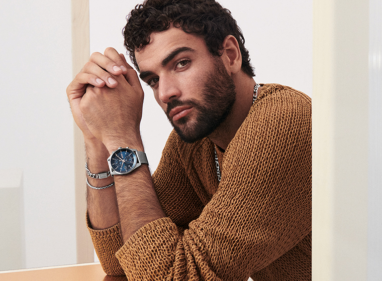 Man in a brown jumper with dark hair and beard wearing a silver watch with a blue dial and steel bracelets  