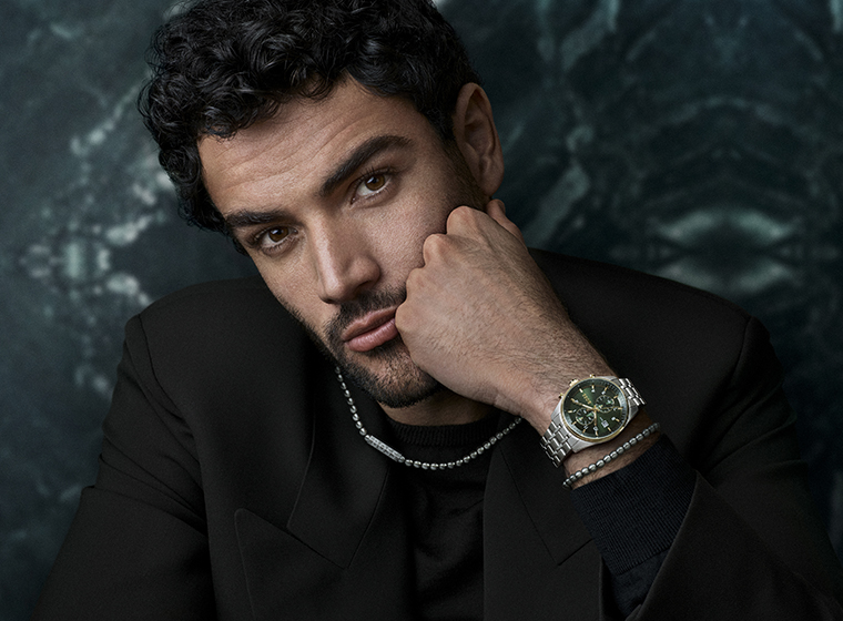 Man in a brown jumper with dark hair and beard wearing a silver watch with a blue dial and steel bracelets  