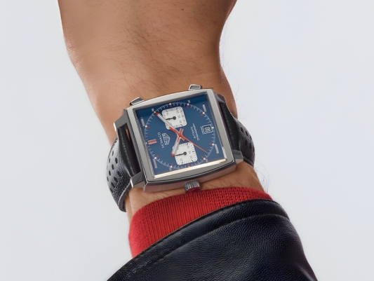 A man wearing a blue dial TAG Heuer watch with a hand on a steering wheel