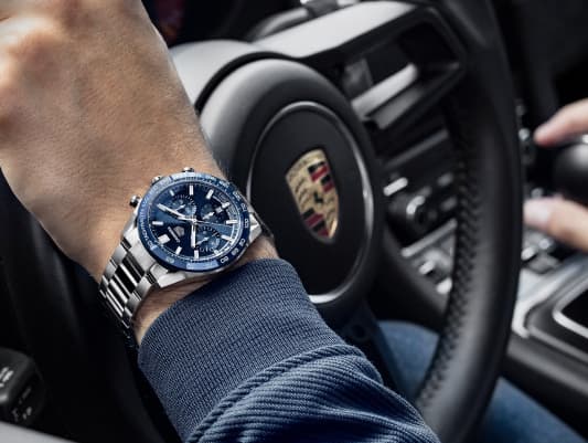 A man wearing a blue dial TAG Heuer watch with a hand on a steering wheel