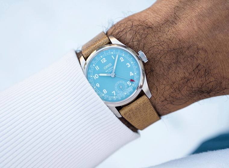 Oris light blue dial watch with a brown strap on a mans wrist wearing a white jumper 