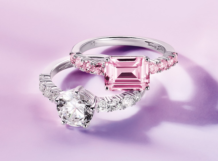 A silver ring with a large pink stone and extra stones on the shoulders laid on top of a silver ring with white stones